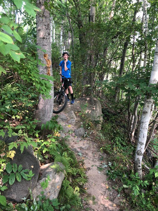A young boy in a blue shirt and helmet stands on a rock in a densely wooded area, holding onto his bike. The trail around him is surrounded by greenery, including trees, ferns, and bushes, creating a natural biking path. Hanson Lake Trail mountain bike trail.