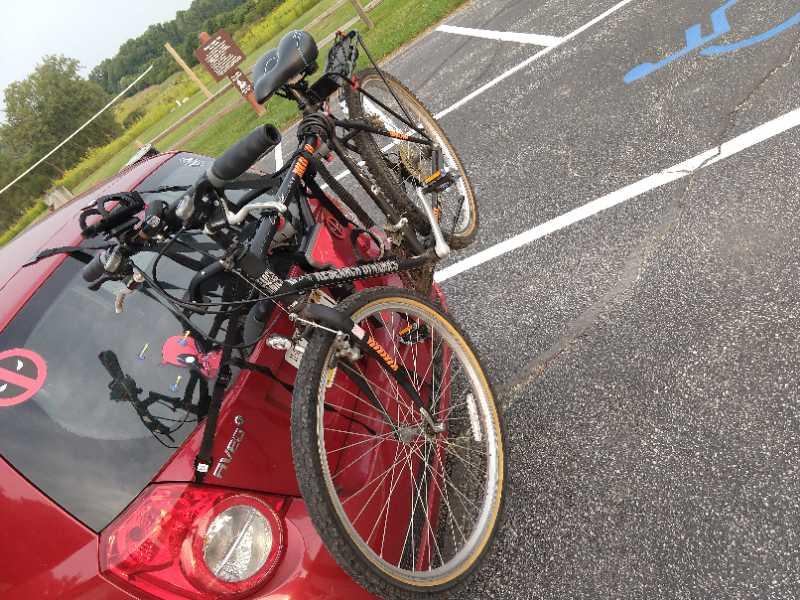 A red car parked in an outdoor setting with two bicycles mounted on a bike rack. The bicycles are positioned upright, displaying various features such as handlebars and tires. In the background, there is a grassy area and a sign post, suggesting a recreational location. Bluhm County Park mountain bike trail.