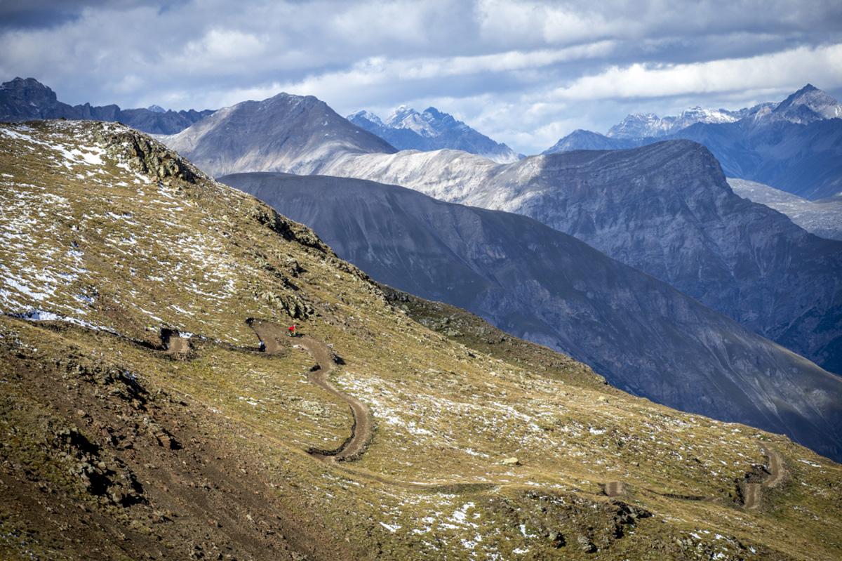 A winding dirt path traverses a mountainous landscape, with hikers visible in the distance. Snow-capped peaks rise majestically in the background under a partly cloudy sky. The terrain is rugged, featuring grassy slopes and rocky outcrops. Coast to Coast mountain bike trail.