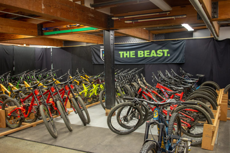A brightly lit bike storage area filled with various mountain bikes arranged neatly. The bikes are predominantly red, green, and black, with some visible details like tires and handlebars. A large black banner reading "THE BEAST." hangs in the background, adding to the cycling atmosphere of the space. The area features wooden supports and a carpeted floor.