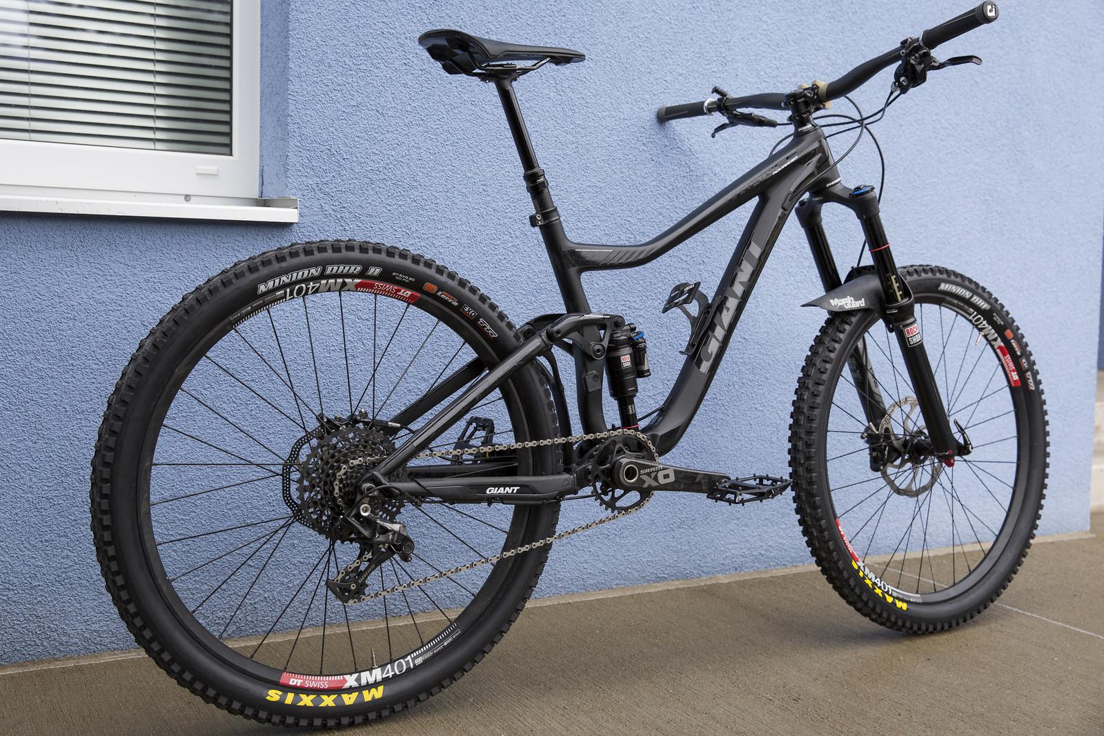 Giant Trance 2: A black mountain bike leaning against a blue wall, featuring wide tires, a sleek frame, and a visible rear suspension system. The bike has a prominent brand logo and is parked on a concrete surface.