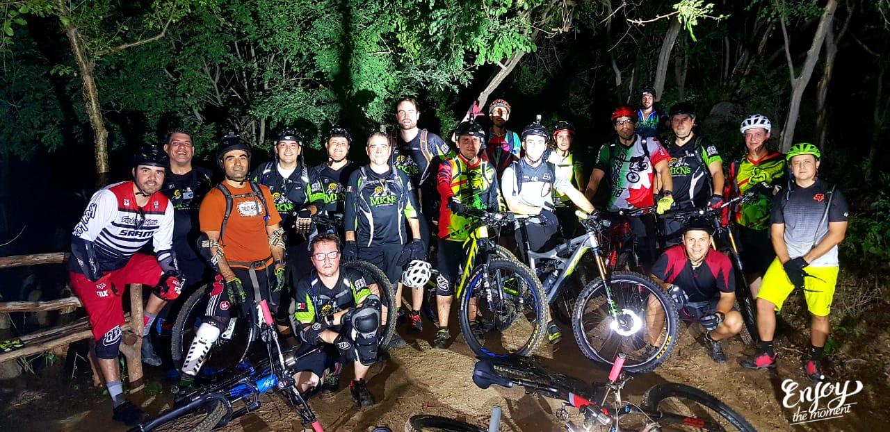 Alt text: A group of mountain bikers posing together at night, surrounded by trees. They are wearing cycling gear and protective equipment, with their bikes in front of them. The scene is illuminated by lights, creating a lively atmosphere. Parque la Colorada mountain bike trail.