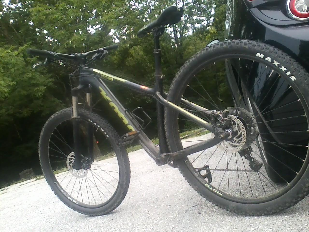 Kona Honzo: A mountain bike with a black and green frame is standing on a gravel surface, partially obscured by a black car. The bike features knobby tires and a disc brake system. Lush green trees are visible in the background, indicating an outdoor setting. The bike appears to be well-used, with dirt visible on the tires and frame.