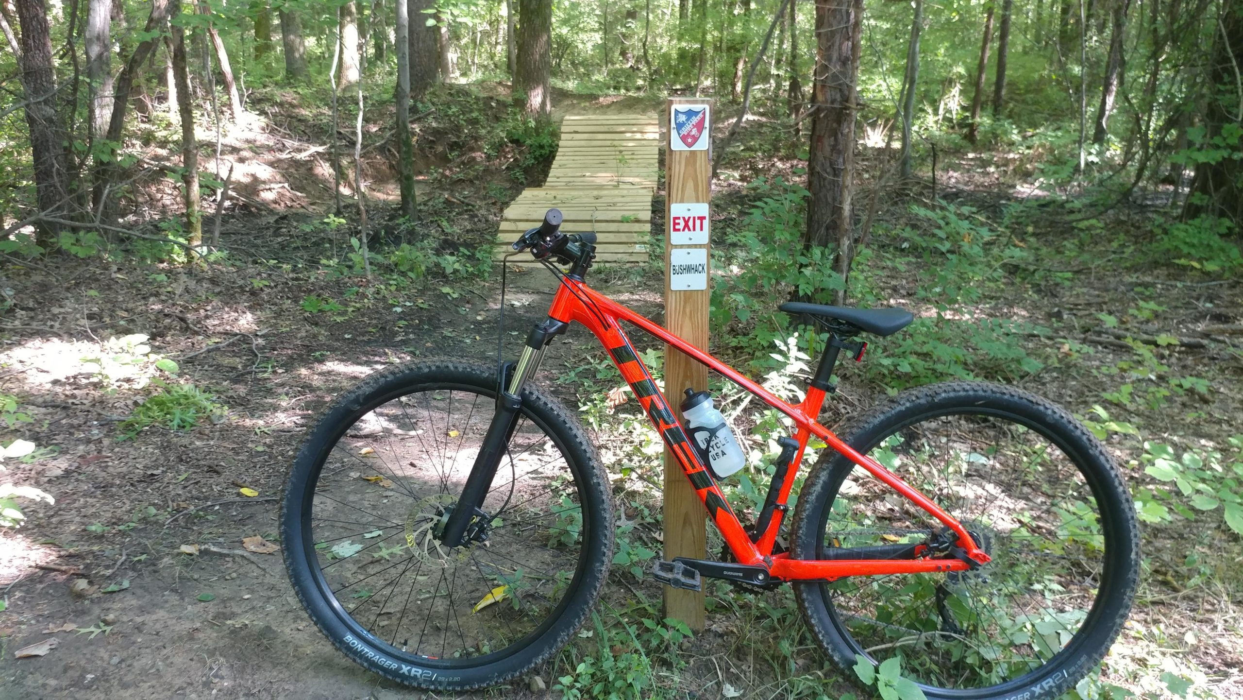 A bright orange mountain bike parked beside a wooden trail exit sign labeled "BUSHWHACK," amidst a lush green forest setting. The bike features thick tires and a water bottle attached to the frame, with sun-dappled light filtering through the trees in the background. Griffin Bike Park mountain bike trail.