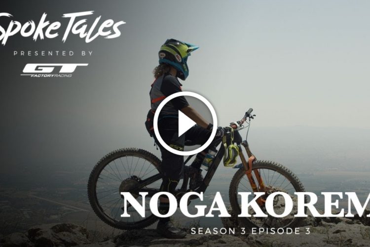 A young mountain biker wearing a helmet and protective gear stands beside their bike, gazing out over a scenic landscape. The image features text that reads "Spoke Tales Presented by GT Factory Racing, Noga Korem, Season 3 Episode 3." A play button icon suggests that it is a video thumbnail.