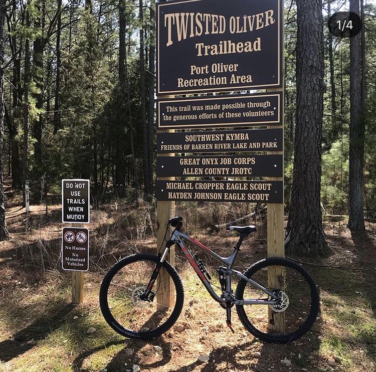 Trek Fuel EX 5 29: A mountain bike is parked next to the Twisted Oliver Trailhead sign at Port Oliver Recreation Area, surrounded by tall pine trees. The sign includes information about the trail and acknowledges the contributions of local volunteers, while nearby signs indicate trail use rules, including a prohibition on horses and motorized vehicles.