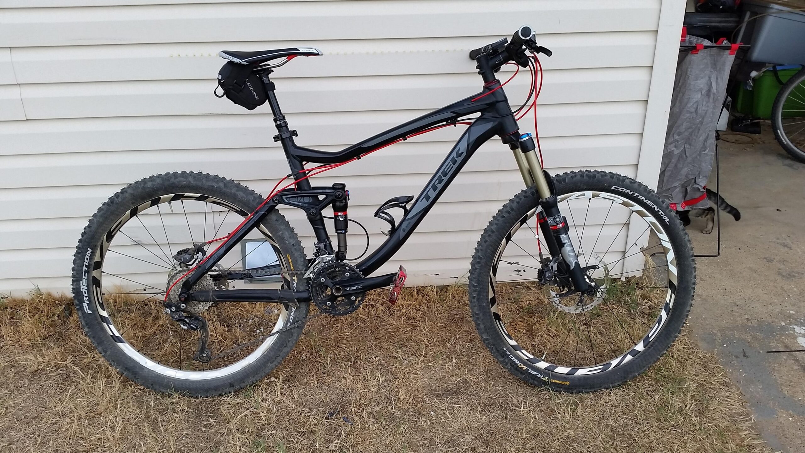 Trek Fuel EX 8: A black and red mountain bike with prominent tires and suspension, positioned against a white wall. The bike features a sleek frame and gear system, with a small bag attached to the seat post. The ground around the bike is grassy, and a small part of a cat can be seen in the background.