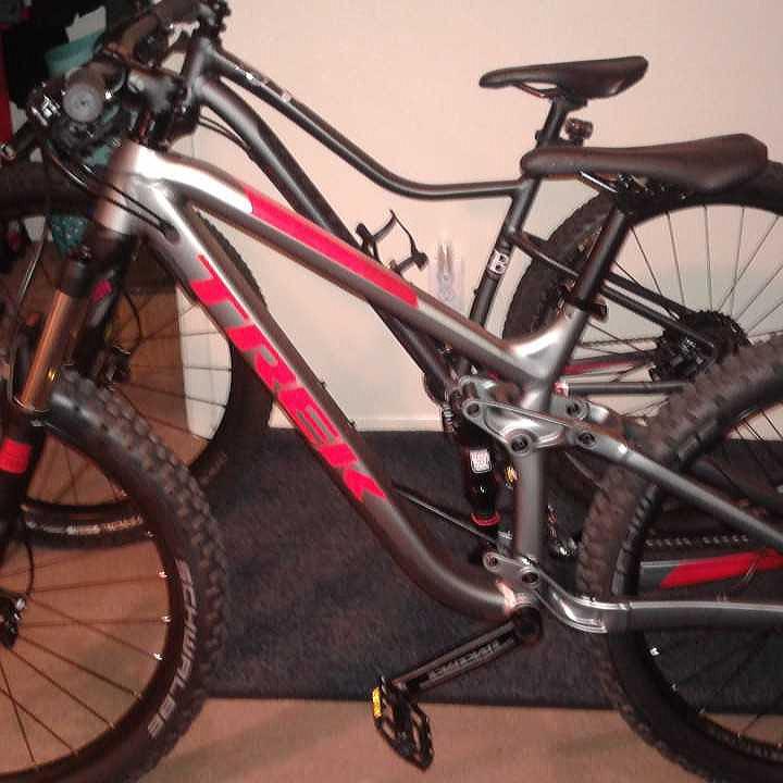 Trek Fuel EX 5: Two mountain bikes are parked closely together in a room. The foreground bike is prominently displaying a silver frame with red accents and the brand name "Trek." The second bike, partially obscured, also has a similar design but is not as clearly visible. Both bikes have thick, rugged tires suitable for off-road terrain. The background shows a plain wall and some indoor items.