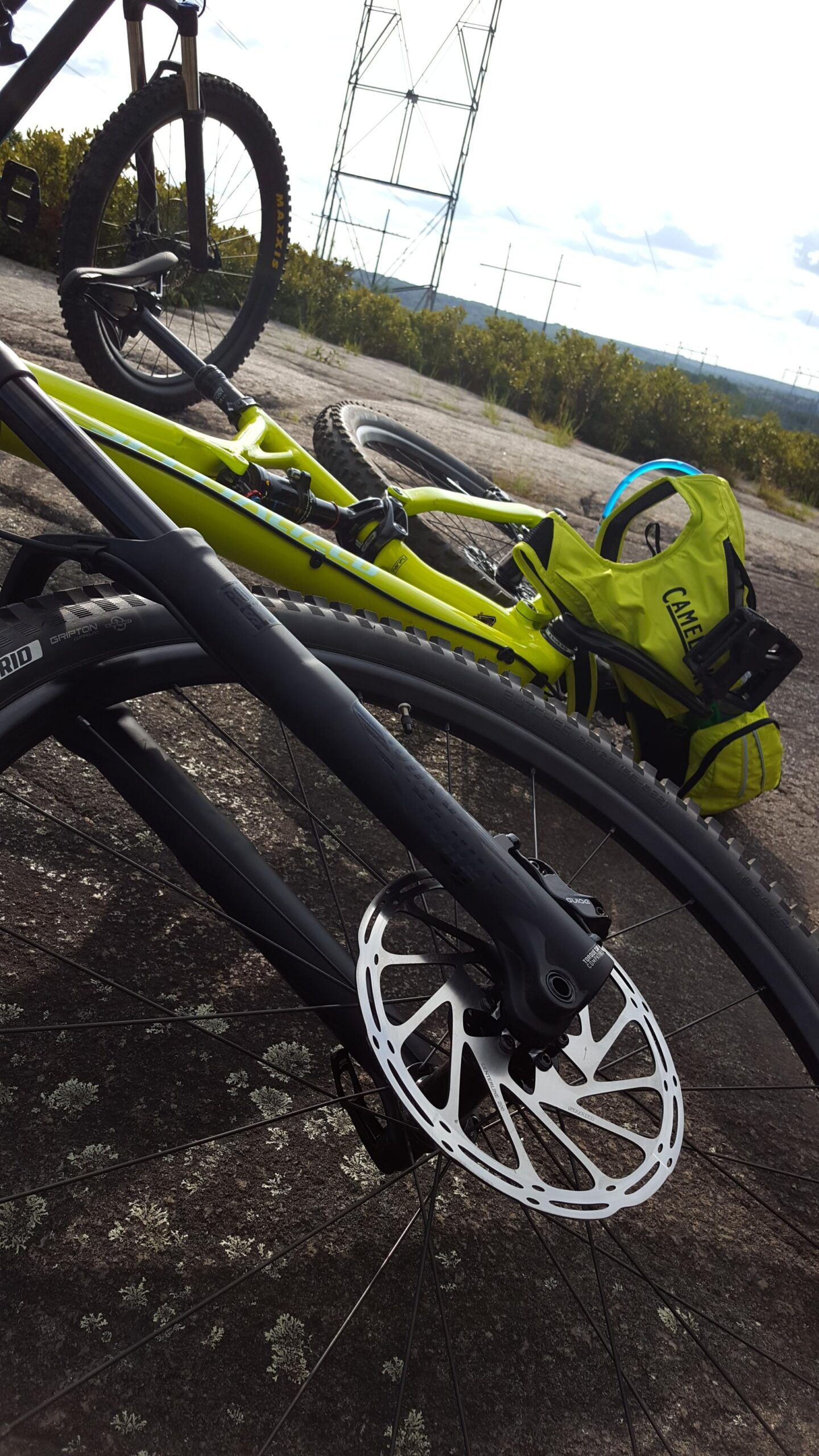Specialized Stumpjumper FSR Comp 29er: Close-up of two mountain bikes resting on a rocky surface, with a focus on the black bike's front wheel and disc brake. In the background, a bright green bike is partially visible along with a yellow hydration pack. The landscape features some greenery and power lines in the distance under a partly cloudy sky.