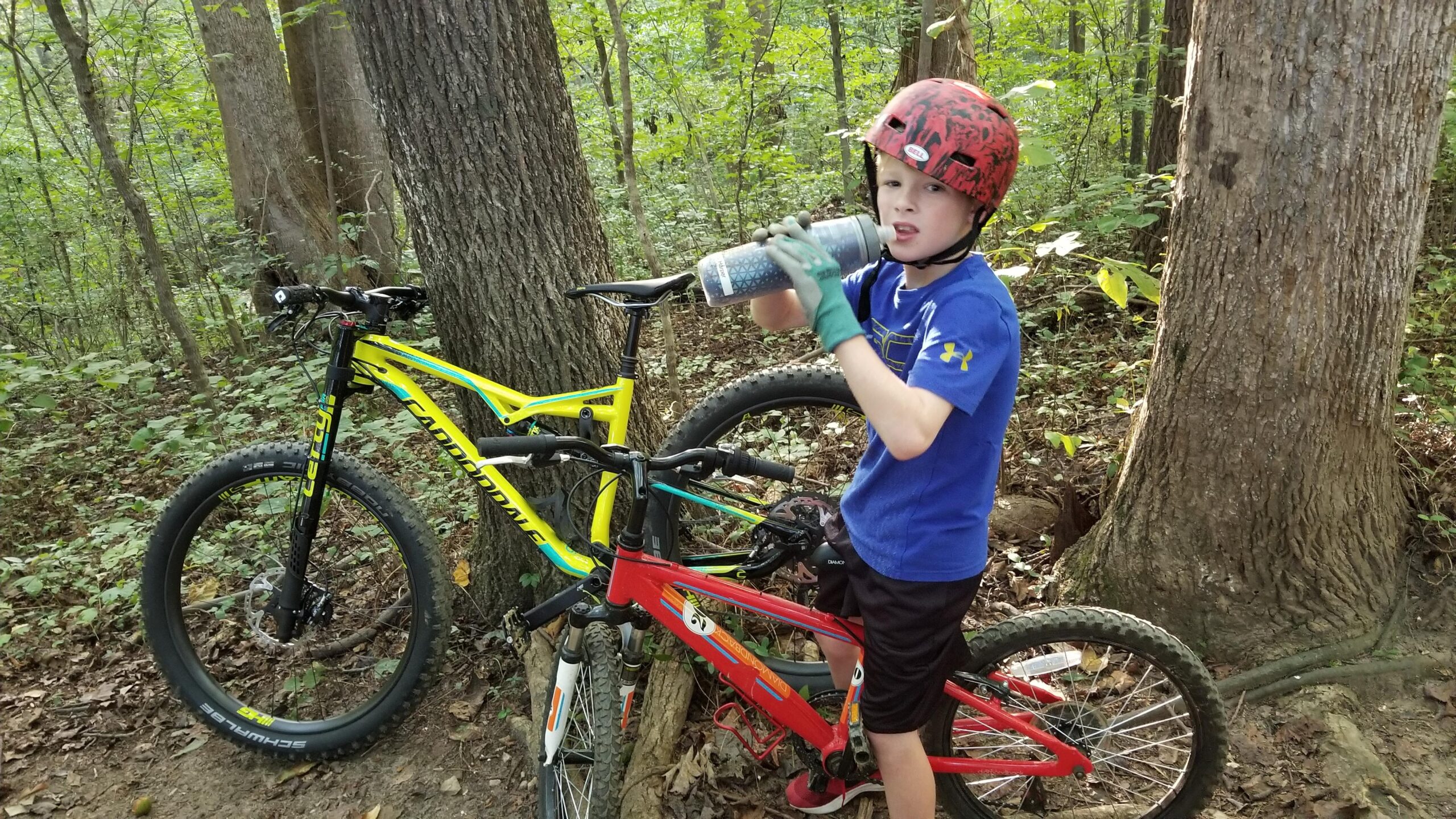 Cannondale Bad Habit: A young boy wearing a red helmet and gloves takes a drink from a water bottle while standing near two mountain bikes in a wooded area. The background features trees and greenery, indicating a natural trail setting. The boy is dressed in a blue shirt and black shorts, with one bike in red and another in yellow.