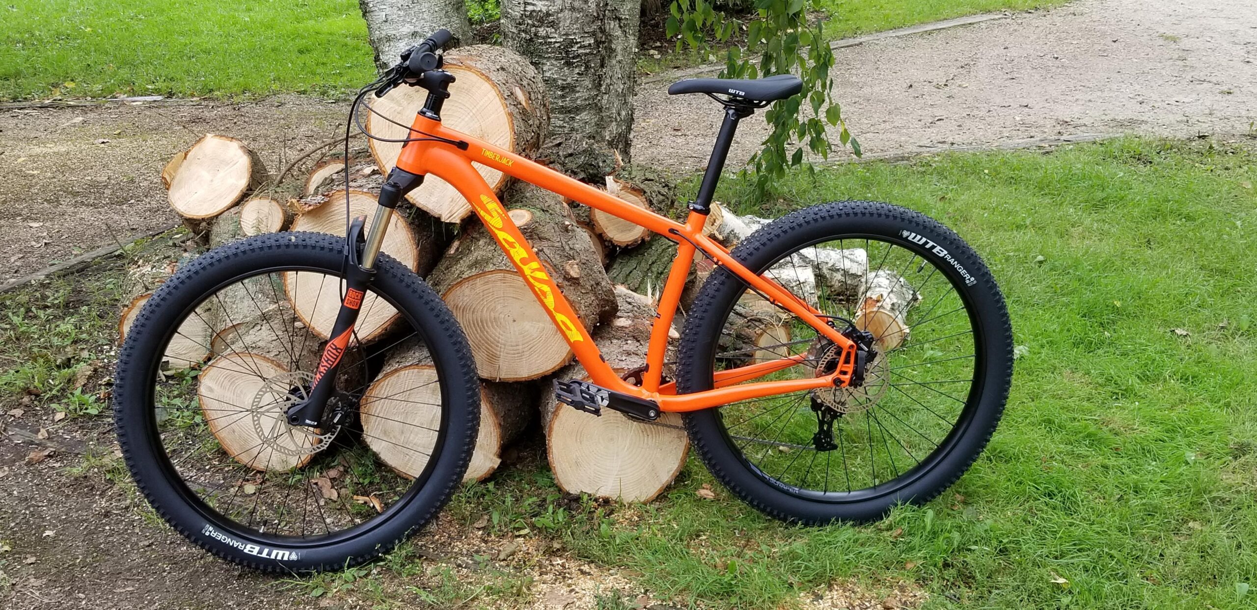 Salsa Timberjack: A bright orange mountain bike is leaning against a stack of cut logs. The bike features thick tires, a sturdy frame, and a front suspension fork. In the background, green grass and part of a gravel path can be seen, indicating an outdoor setting.