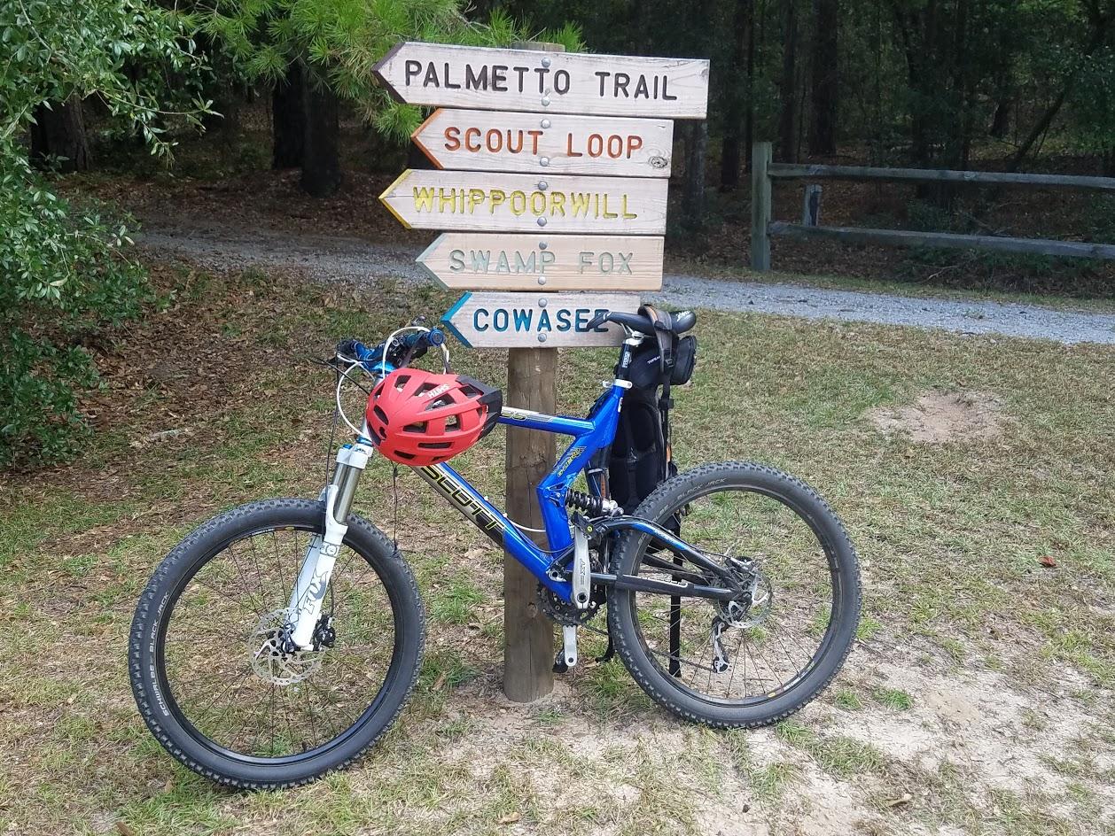 A blue mountain bike with a red helmet rests next to a wooden trail signpost indicating directions for various trails, including Palmetto Trail, Scout Loop, Whippoorwill, Swamp Fox, and Cowasee, set in a wooded area. Poinsett State Park mountain bike trail.