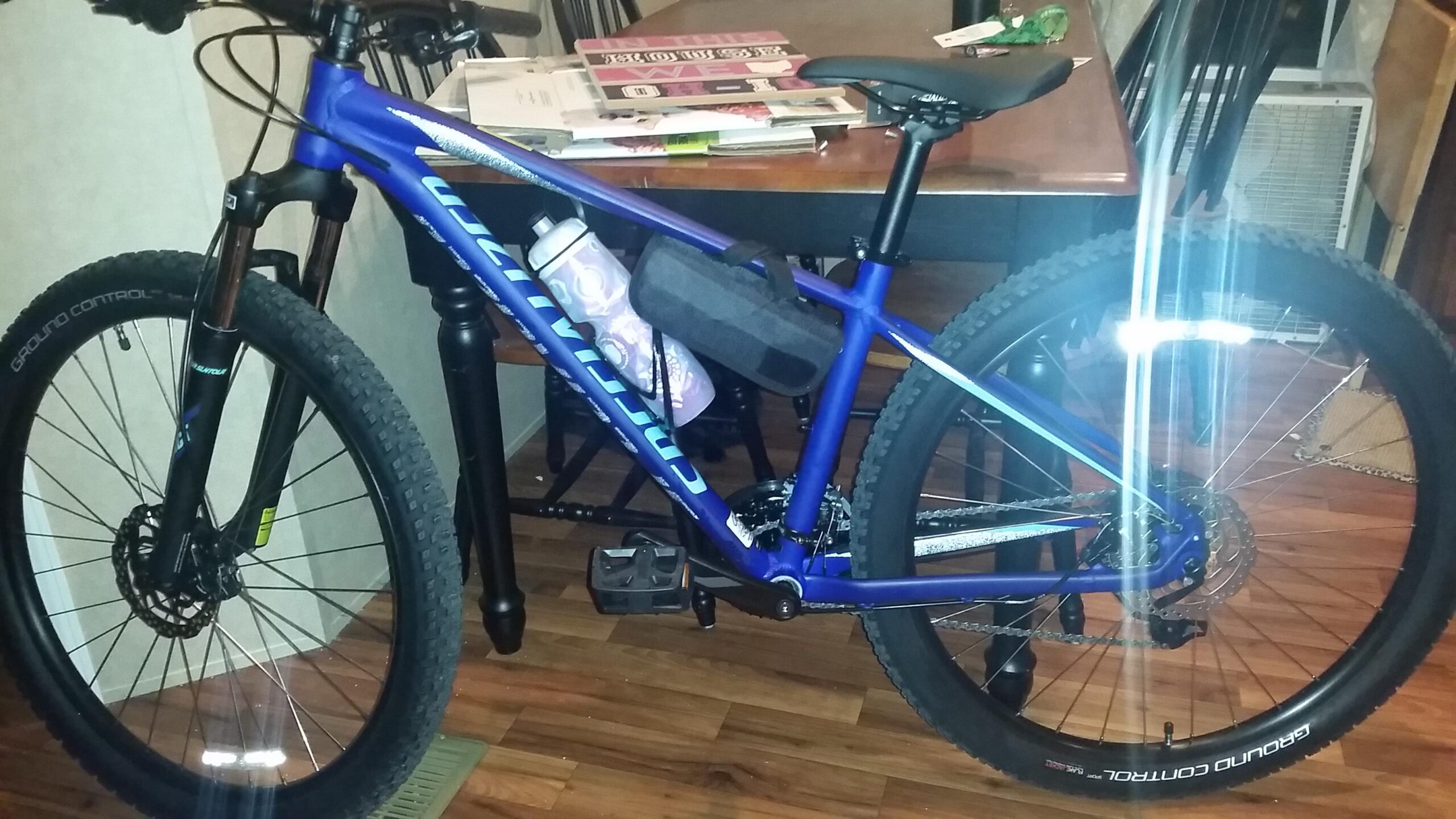 Specialized Pitch: A blue mountain bike is positioned indoors, leaning against a table. The bike features wide tires labeled "Ground Control," a water bottle attached to the frame, and a small black bag on the side. In the background, there are stacks of papers and furniture, with a hint of a cooling unit visible. The wooden floor adds warmth to the scene.