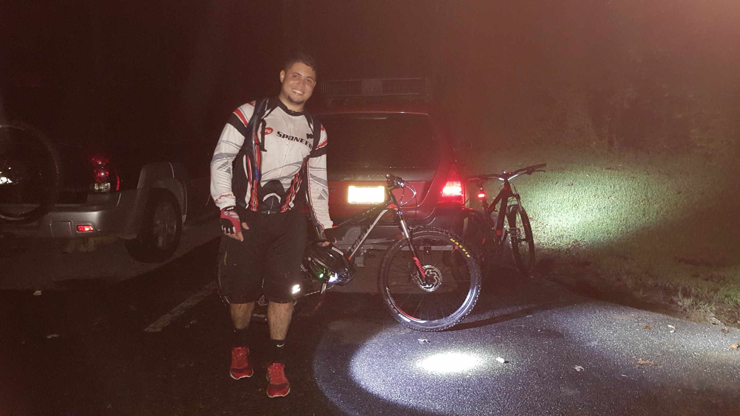 A cyclist wearing a sport jersey and shorts stands next to a parked car, holding a helmet. Two mountain bikes are propped against the car, illuminated by a spotlight on the ground. The setting appears to be dark, likely during nighttime, with grass and trees visible in the background. Chimney Rock mountain bike trail.