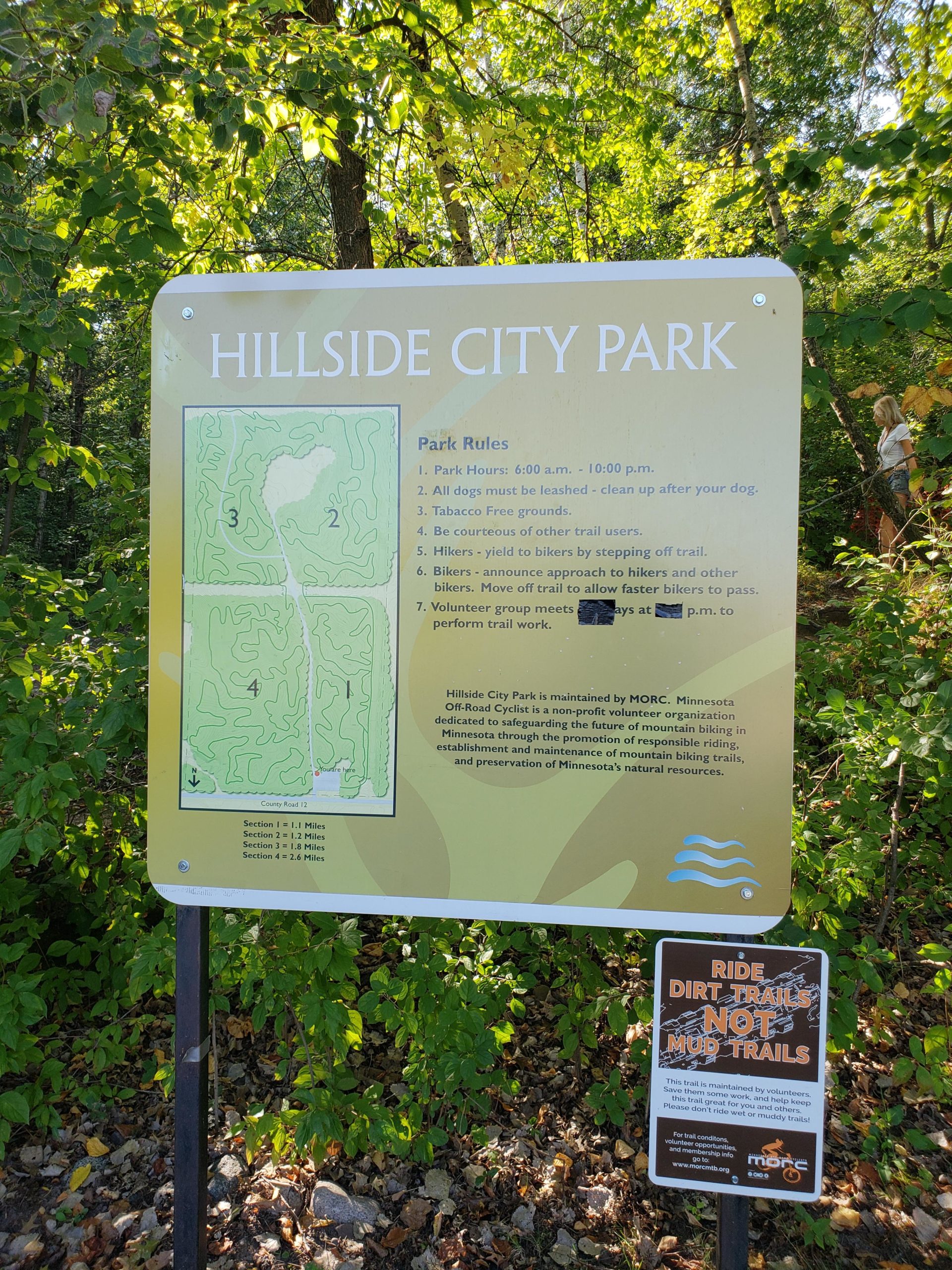 Signage for Hillside City Park featuring park rules, hours, and a trail map. The sign emphasizes rules for dog owners, bikers, and hikers, and is surrounded by green foliage. An additional sign below advises users to ride on dirt trails, not muddy ones. Hillside Park mountain bike trail.