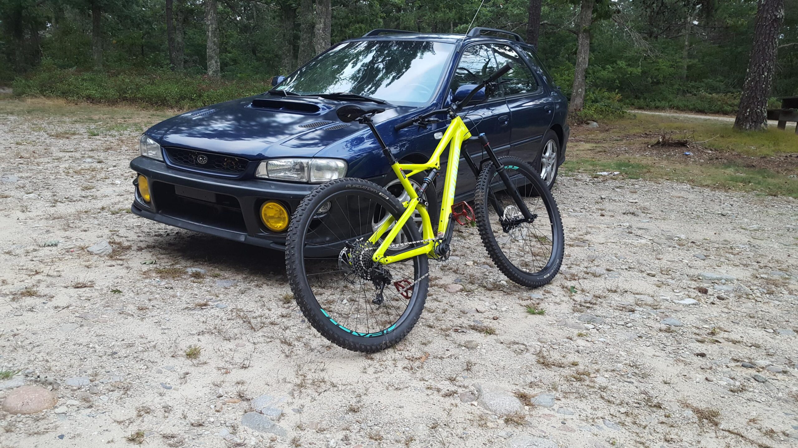 Specialized Stumpjumper FSR Comp 29: A bright yellow mountain bike is leaned against a dark blue car parked on a gravel surface, surrounded by trees in a natural setting. The car features a subtle sporty design with yellow fog lights, and both the bike and car suggest an outdoor adventure theme.