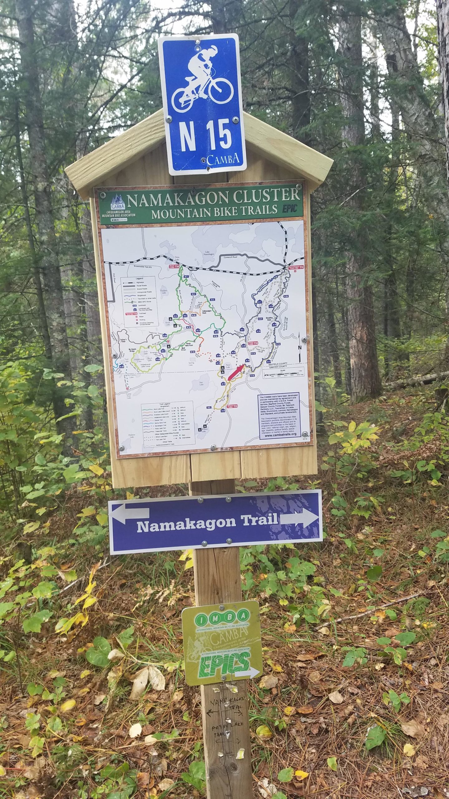 A trail sign displaying a map of the Namakagon Cluster Mountain Bike Trails, featuring trail markers and directions. The sign includes a blue bike symbol and indicates the location of the Namakagon Trail, surrounded by trees and foliage in a forested area. Patsy Lake Trail mountain bike trail.