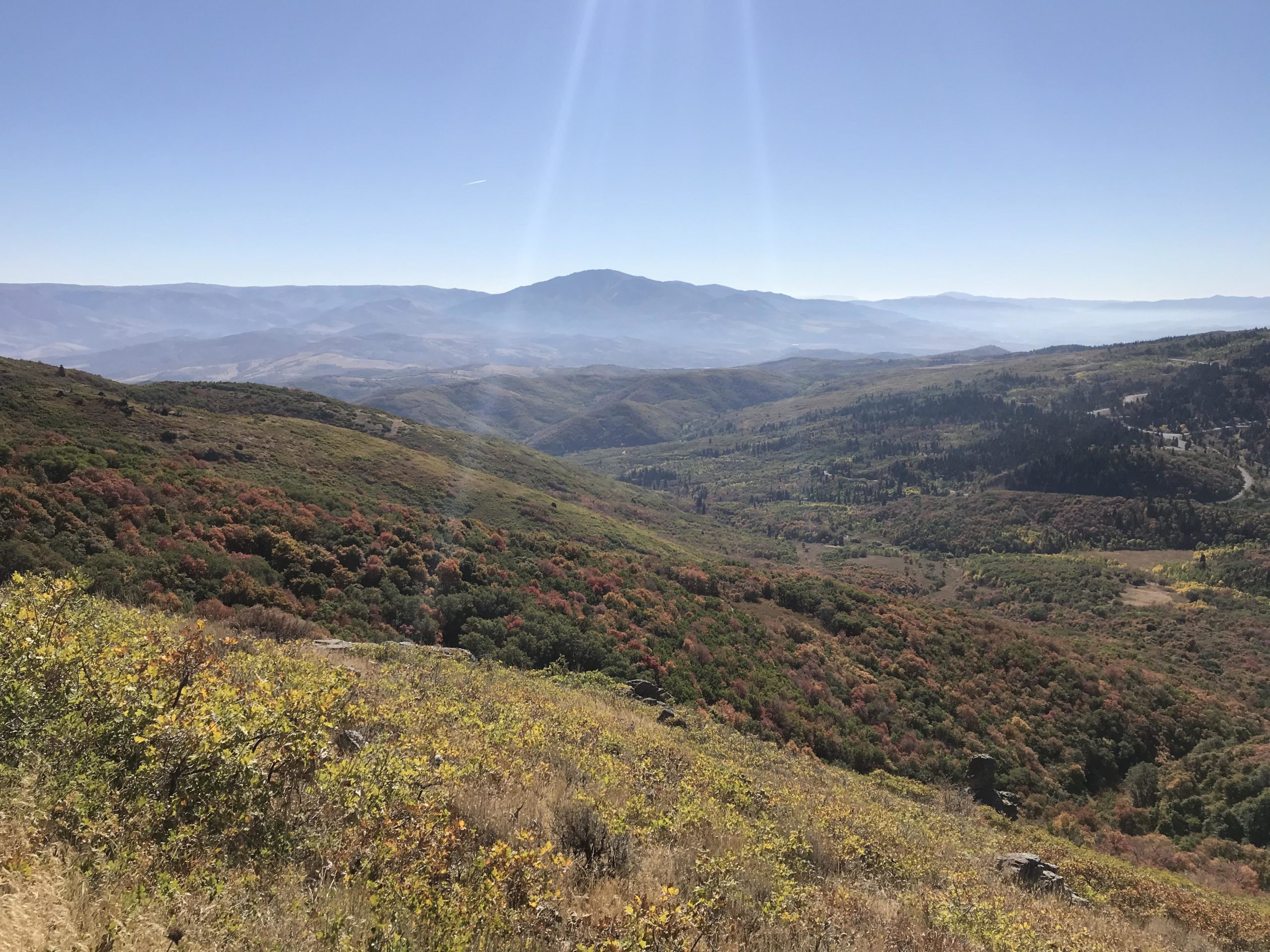 A panoramic view of rolling hills and mountains under a clear blue sky, showcasing vibrant autumn foliage in varying shades of green, orange, and red. The landscape is dotted with patches of trees and distant valleys, with soft morning light illuminating the scene. Snowbasin Resort mountain bike trail.