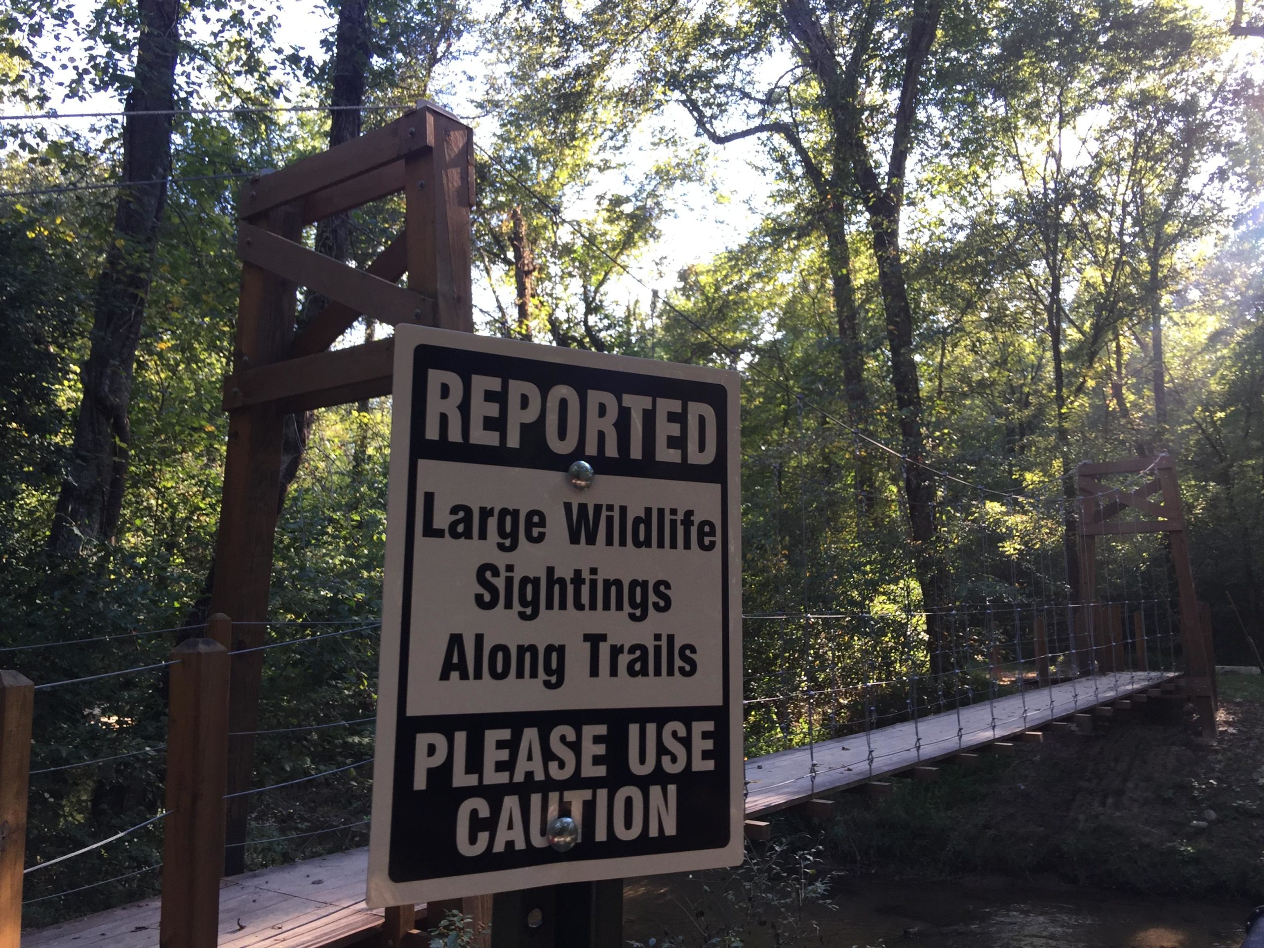 Sign warning about large wildlife sightings along trails, with a wooden bridge visible in the background, surrounded by trees and natural scenery. Cub Creek Trails mountain bike trail.