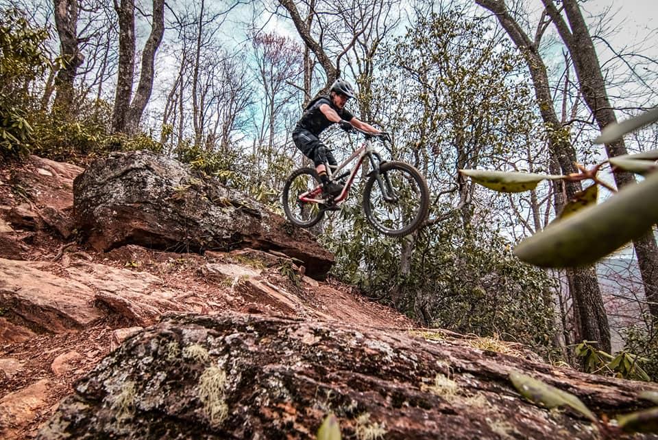 YT Industries YT Jeffsy CF: A mountain biker performing a jump off a rocky ledge on a forest trail, surrounded by trees and greenery. The cyclist is wearing a helmet and is captured in mid-air, showcasing an action-packed moment in off-road biking.