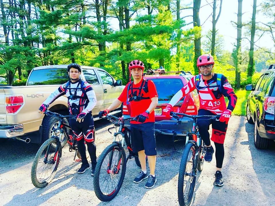 Three mountain bikers stand posed beside their bicycles in a wooded outdoor setting. They are wearing colorful biking gear, including helmets, and are positioned near parked vehicles. The background features trees and a sunny sky, indicating a pleasant day for biking. Six Mile Run mountain bike trail.