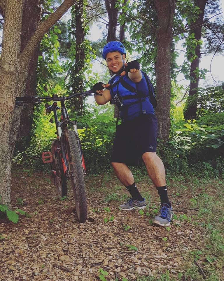 A person wearing a blue helmet and matching shirt is smiling and posing beside a mountain bike in a wooded area. They are standing on a bed of leaves and the surrounding trees are lush with greenery. Six Mile Run mountain bike trail.