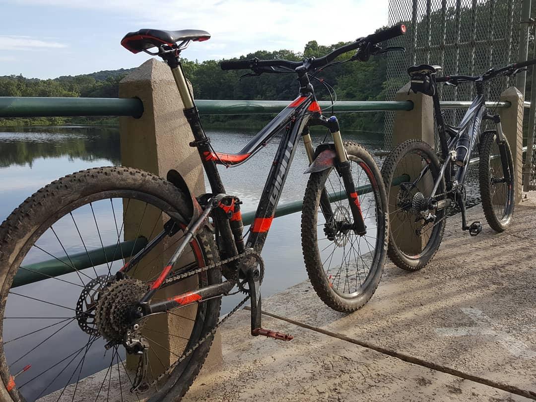 Two mountain bikes are parked on a concrete surface next to a calm river. The bike on the left has a red and black design, while the bike on the right is mostly black. There is a green railing and trees in the background, under a clear blue sky. The setting suggests a scenic outdoor area ideal for cycling. Chimney Rock mountain bike trail.