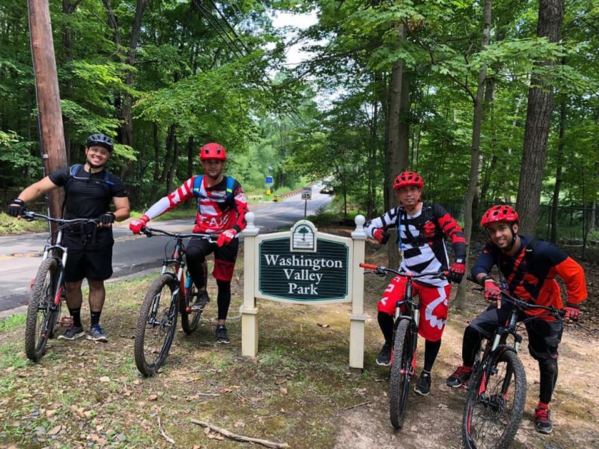 Four cyclists in biking gear pose in front of a sign that reads "Washington Valley Park." The group includes two men wearing red helmets and colorful jerseys, while standing beside their mountain bikes. Lush green trees surround the area, and a paved road is visible in the background. Chimney Rock mountain bike trail.