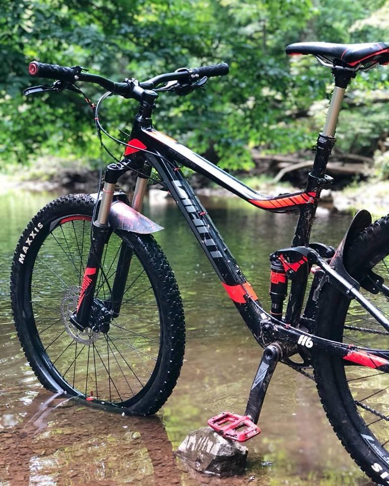 Giant Stance 27.5 2: Mountain bike partially submerged in shallow water, surrounded by lush greenery. The bike has a black and red color scheme, with visible tires and pedals resting on a rock.