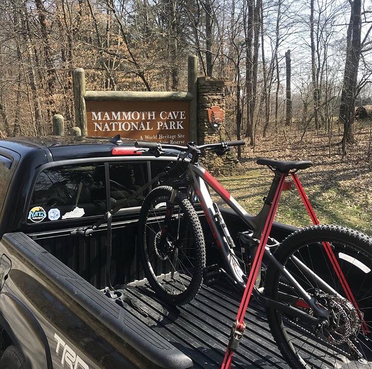 Trek Fuel EX 5 29: A pickup truck parked at Mammoth Cave National Park, featuring a bicycle in the truck bed. The background includes a wooden park sign indicating the park's name and its status as a World Heritage Site, surrounded by trees in a wooded area.