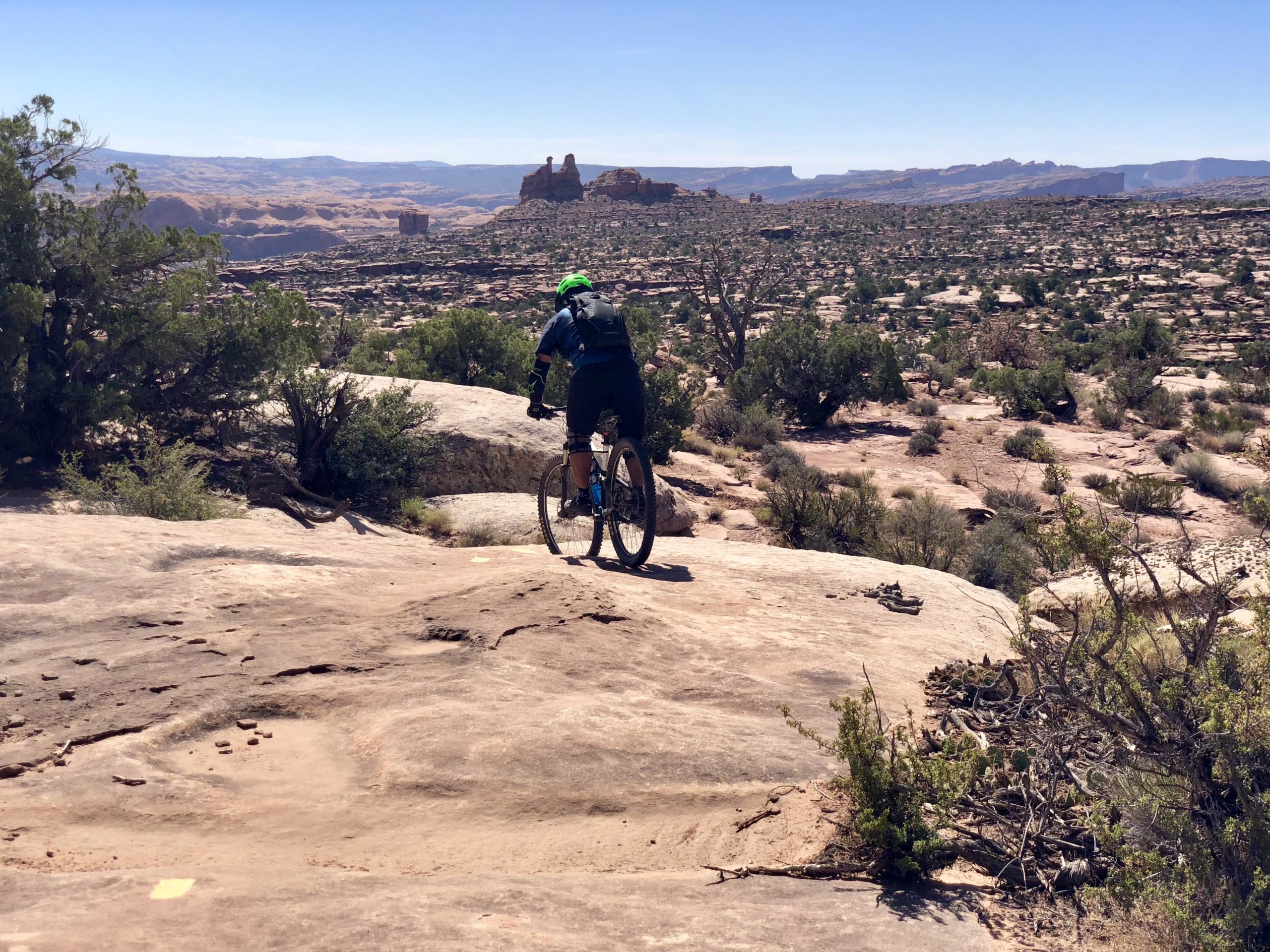 A mountain biker rides over rocky terrain while overlooking a vast landscape with mesas and distant mountains under a clear blue sky. Vegetation, including shrubs and small trees, is scattered throughout the foreground. Gold Bar Rim mountain bike trail.