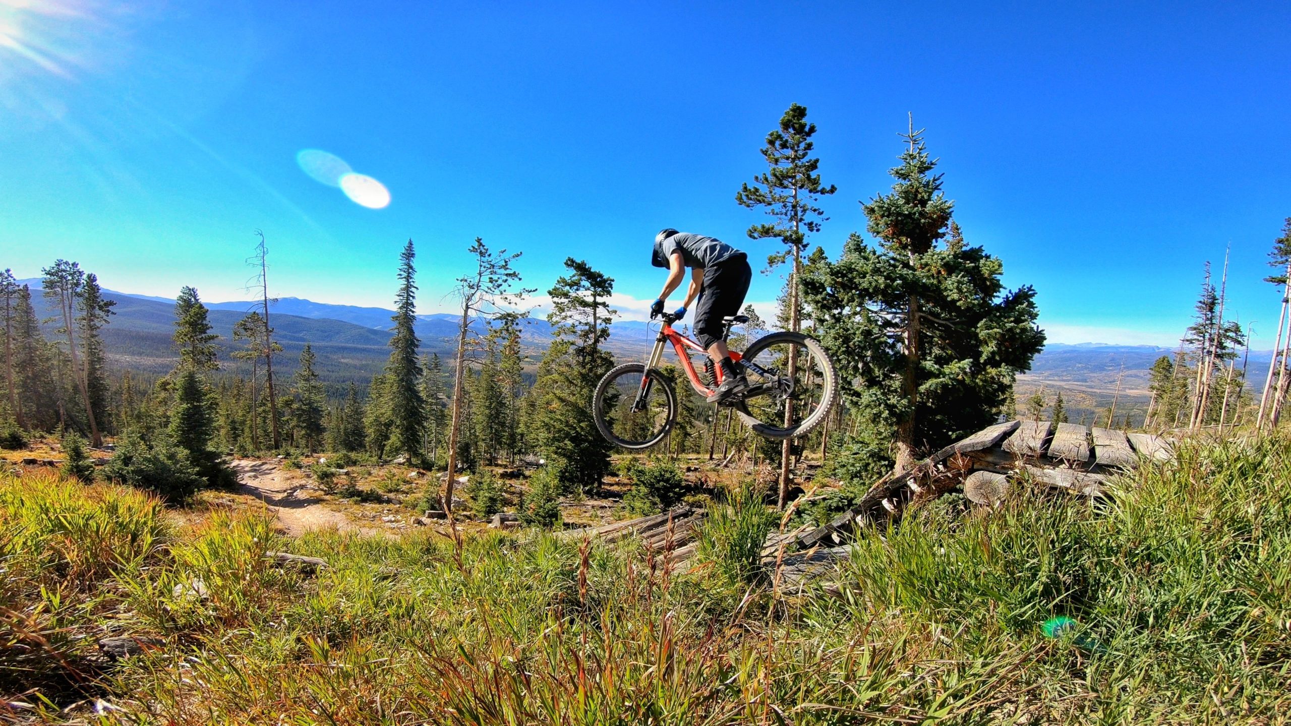 A mountain biker performing a jump over a wooden obstacle on a trail surrounded by tall trees and mountain scenery under a clear blue sky. Trestle Bike Park mountain bike trail.