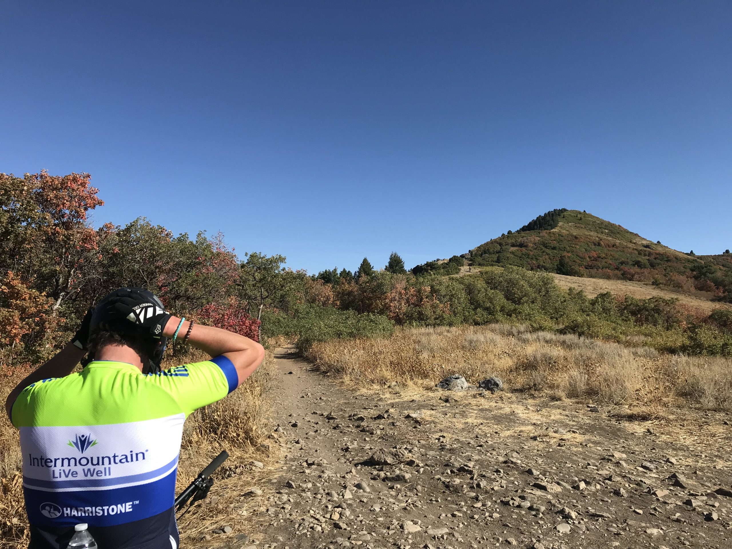 A mountain biker wearing a bright green and blue cycling jersey stands on a rugged trail. The landscape features vibrant autumn foliage, with trees displaying shades of red and orange. In the background, a green hill rises under a clear blue sky. The biker is adjusting their helmet, preparing for the ride ahead. Snowbasin Resort mountain bike trail.