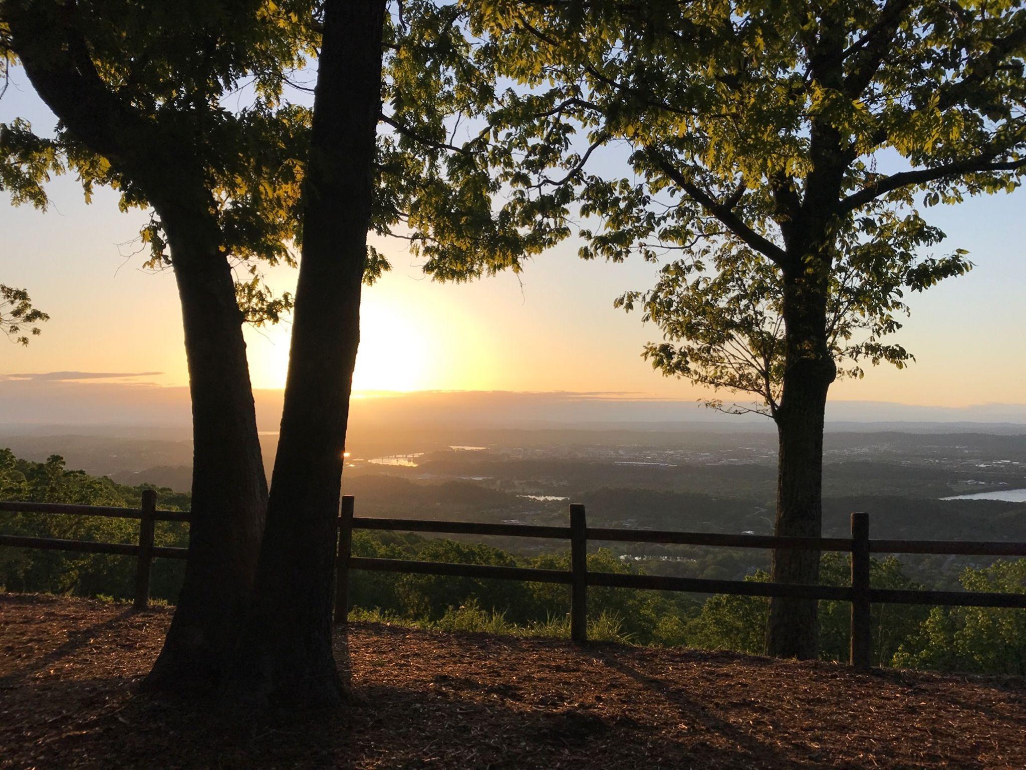 Sunset view from a hillside, framed by two trees. The horizon displays a mix of warm orange and cool blue tones, with distant hills and water bodies visible below. A wooden fence lines the foreground, set against a backdrop of lush greenery. Raccoon Mountain Trail Network mountain bike trail.