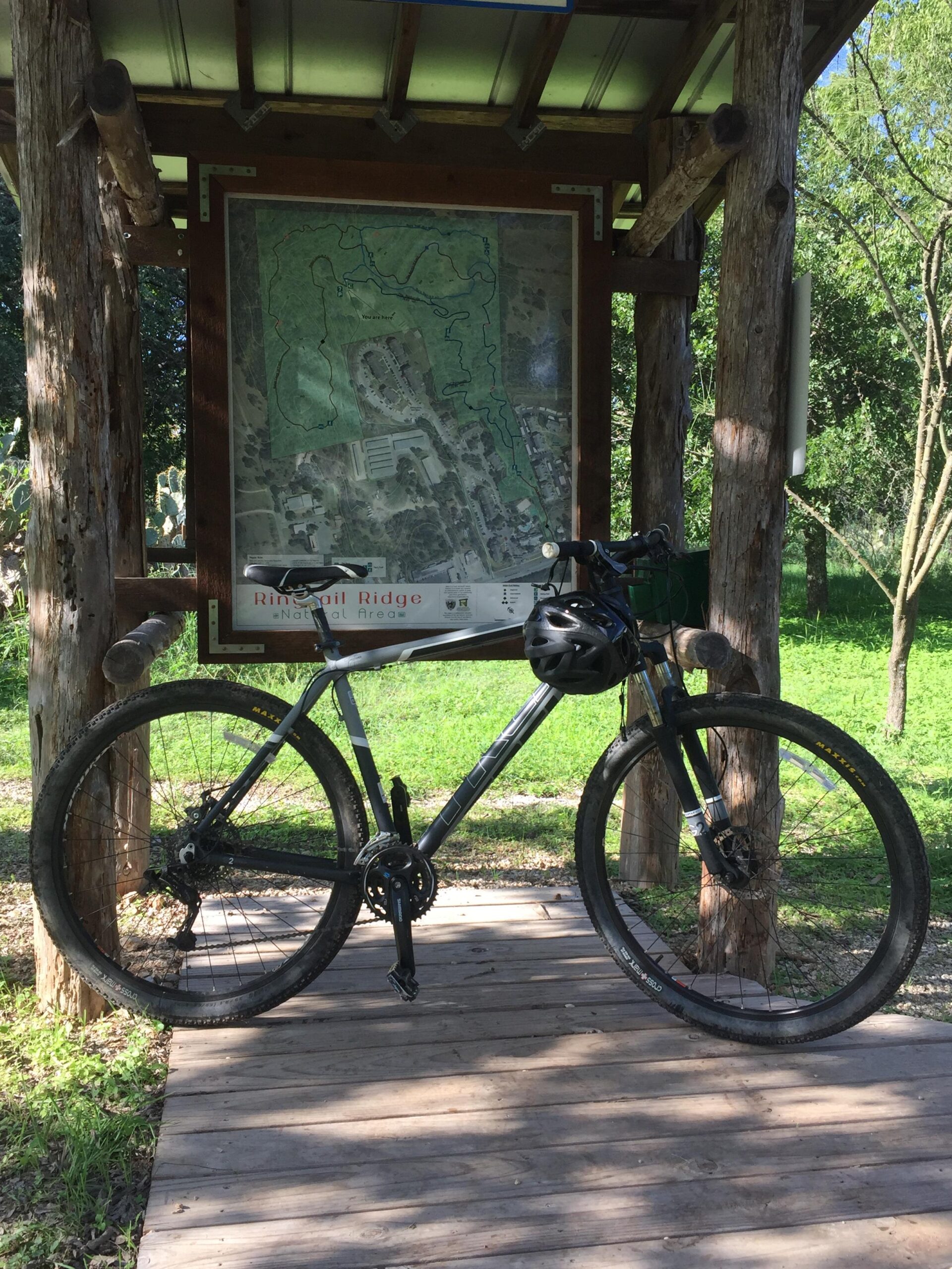 Trek Trek Marlin 4 (29er): A mountain bike parked on a wooden platform in front of a map display at a natural area. The map details trails and features of Ringtail Ridge. Surrounding greenery and trees offer a scenic natural backdrop.
