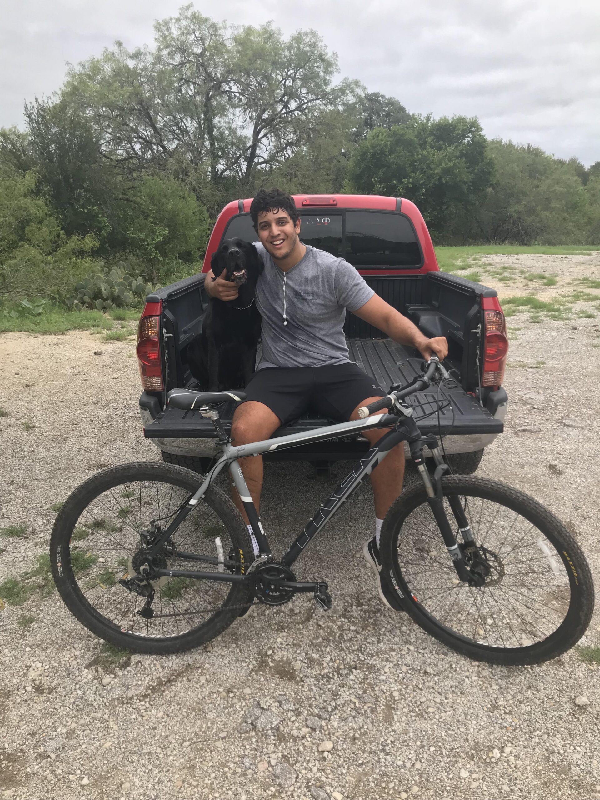 Trek Trek Marlin 4 (29er): A young man sitting in the back of a pickup truck, smiling and posing with a black dog. He is wearing a gray t-shirt and shorts, with a mountain bike resting beside him. The background features a natural landscape with trees and a cloudy sky.