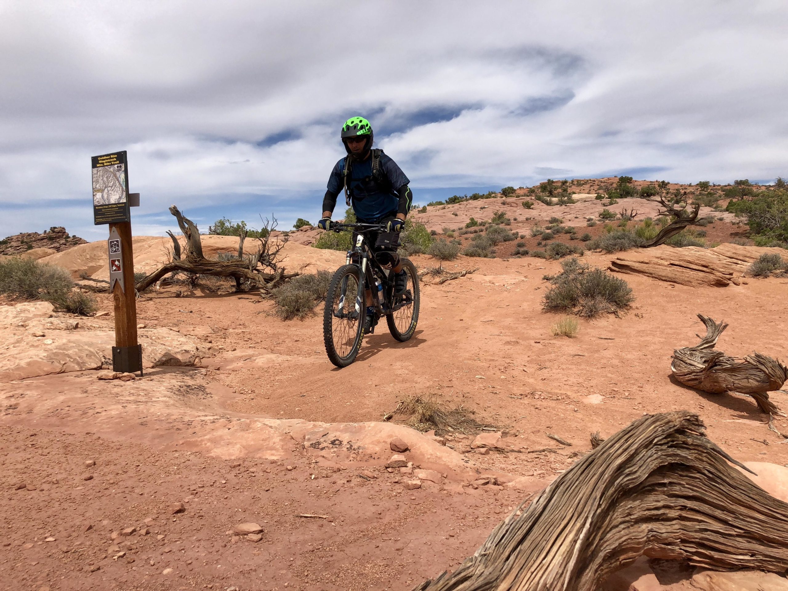 A mountain biker wearing a green helmet and blue jacket rides on a rocky, dirt trail surrounded by sparse vegetation and rock formations, with a trail sign in the background indicating directions. The sky is partly cloudy, suggesting an outdoor setting. Gold Bar Rim mountain bike trail.