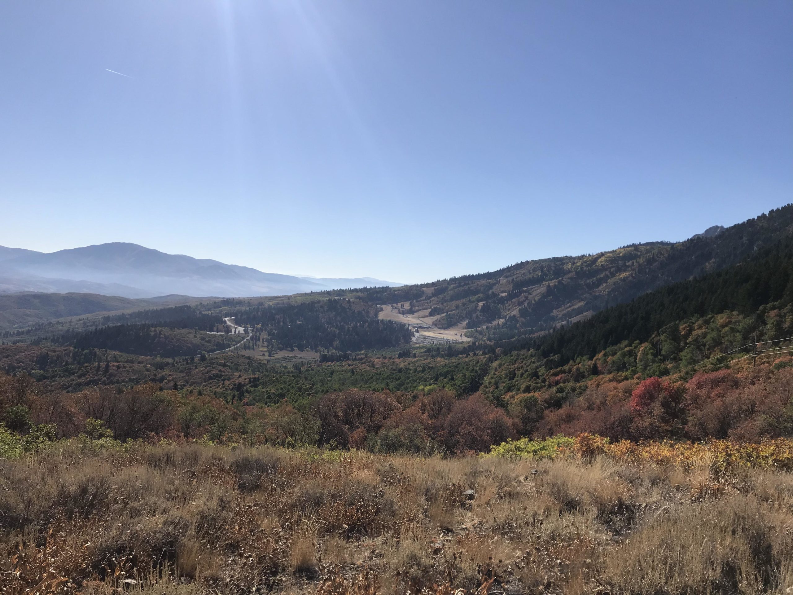 A panoramic view of rolling hills and mountains under a clear blue sky. The landscape showcases a mix of greenery and autumn foliage, with vibrant reds, oranges, and yellows. A winding road can be seen cutting through the valley, surrounded by trees and shrubs. Snowbasin Resort mountain bike trail.