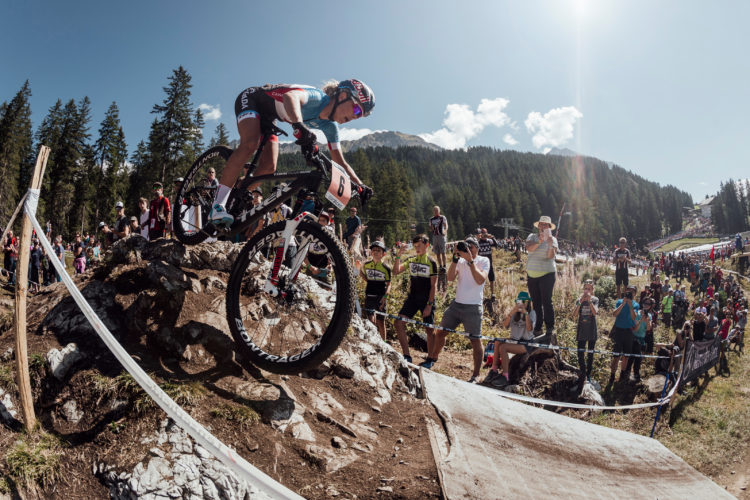 A mountain biker descends a rocky trail during a race, showcasing skill and agility. Spectators line the course, capturing the moment with their cameras as the sun shines brightly overhead, surrounded by forested hills.