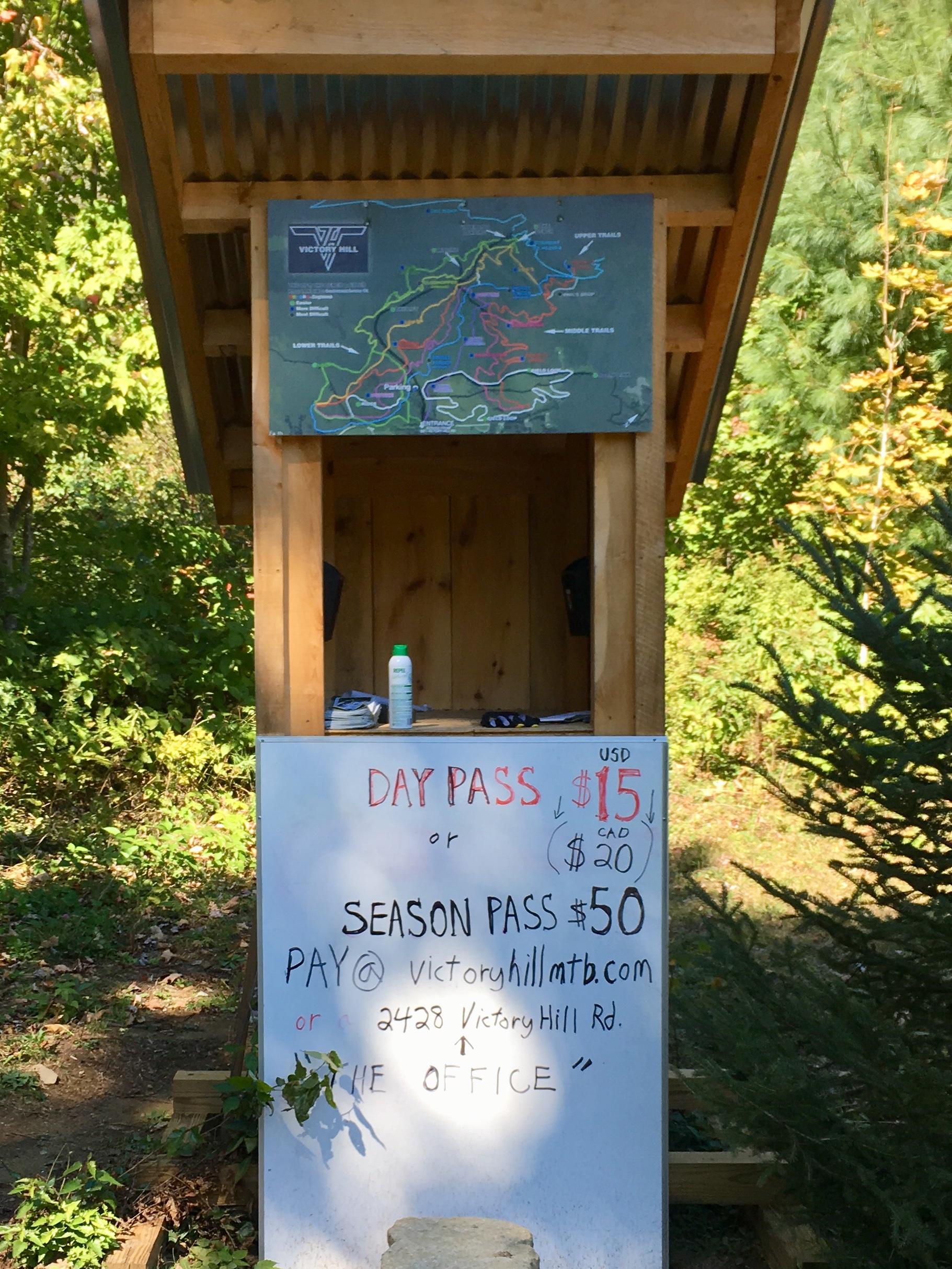 Alt text: A wooden kiosk displaying a trail map and pricing for mountain biking passes. The sign lists a day pass for $15 USD or $20 CAD, and a season pass for $50, with instructions for payment and location details. Surrounding vegetation is visible in the background. Victory Hill Sector mountain bike trail.