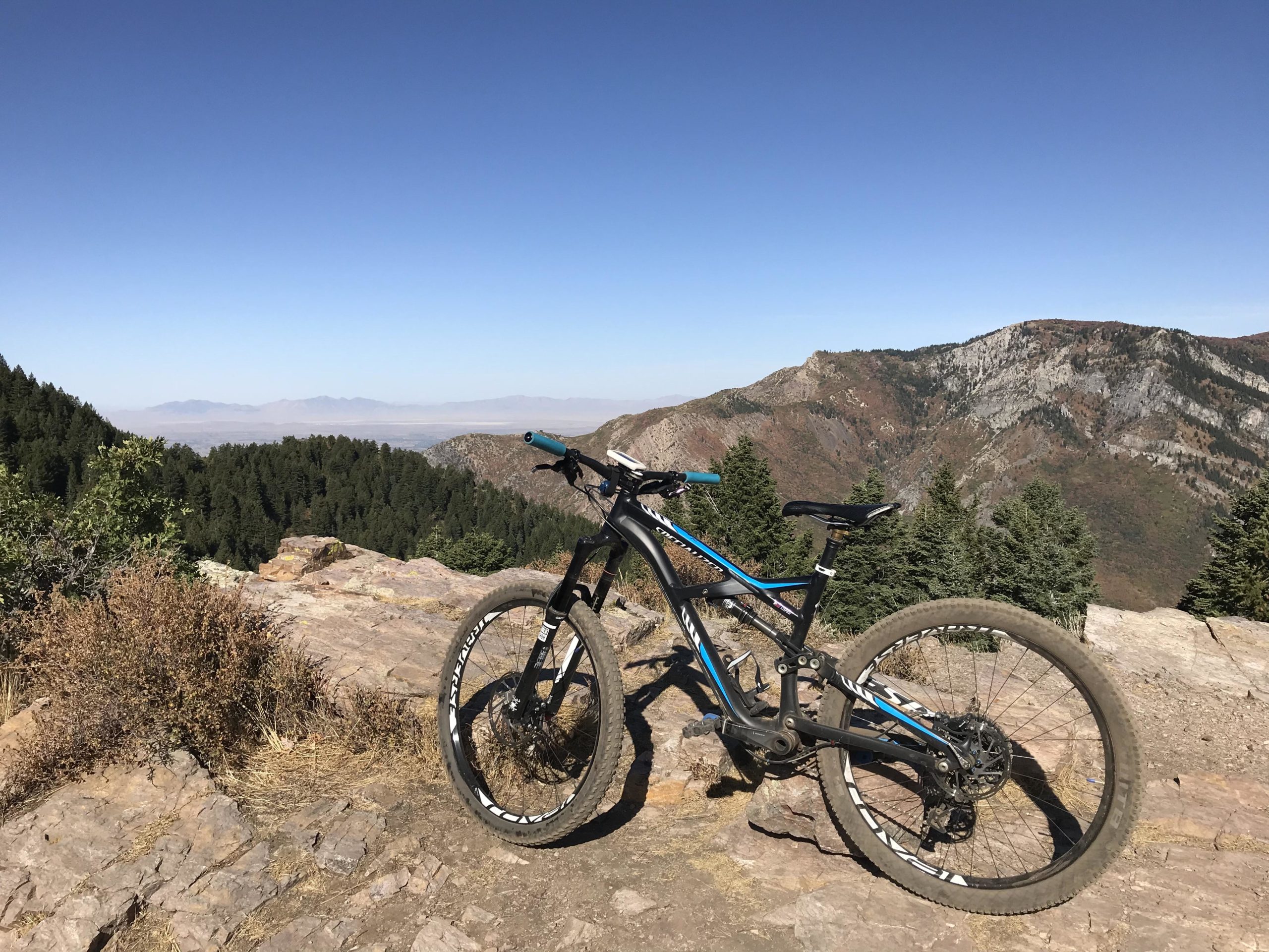 A mountain bike rests on rocky terrain with a panoramic view of mountains and valleys in the background, under a clear blue sky. Lush green trees are visible in the foreground, showcasing a scenic outdoor adventure location. Snowbasin Resort mountain bike trail.