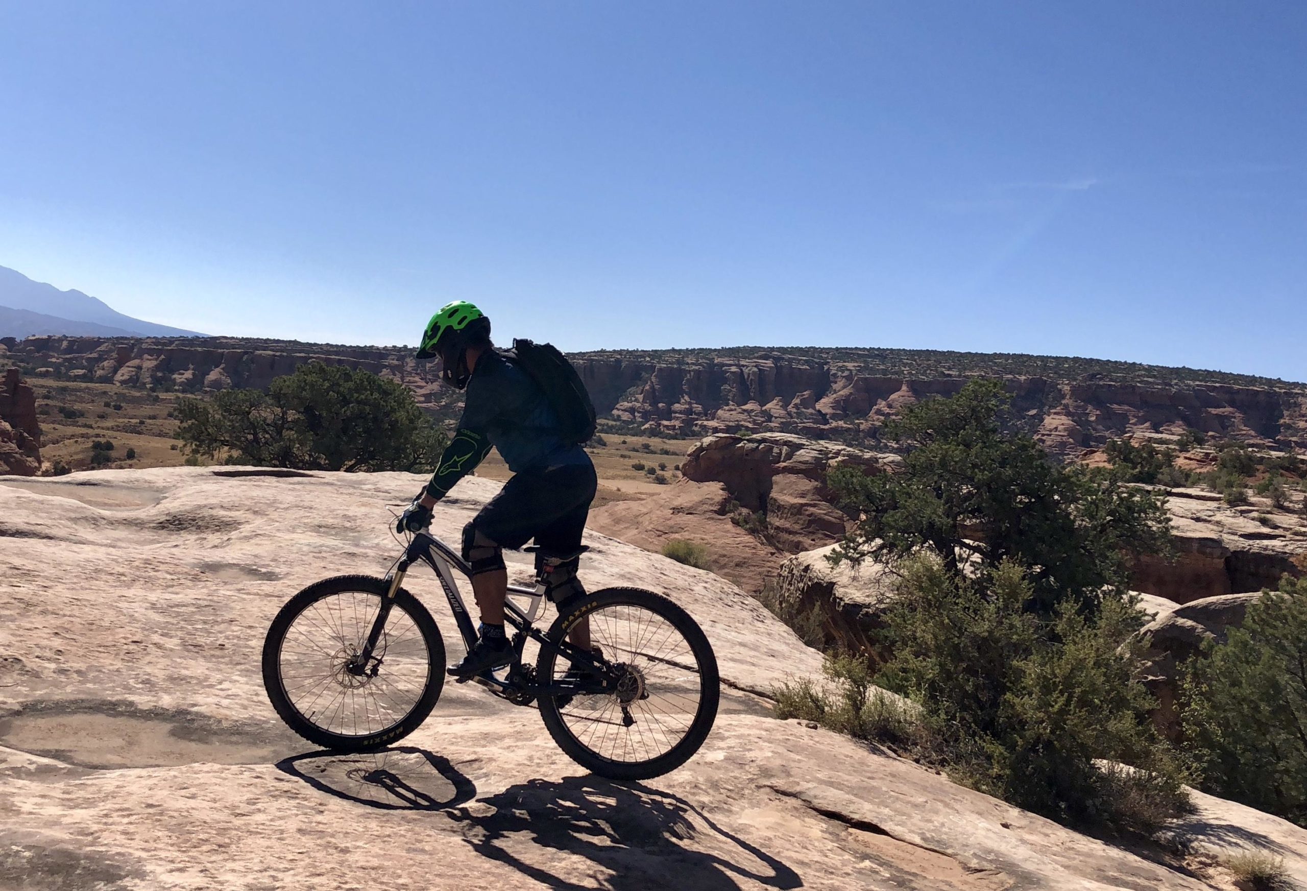A mountain biker riding along a rocky terrain with a scenic landscape in the background, featuring cliffs and vegetation under a clear blue sky. The cyclist is wearing a bright green helmet and protective gear, navigating the challenging outdoor environment. Gold Bar Rim mountain bike trail.