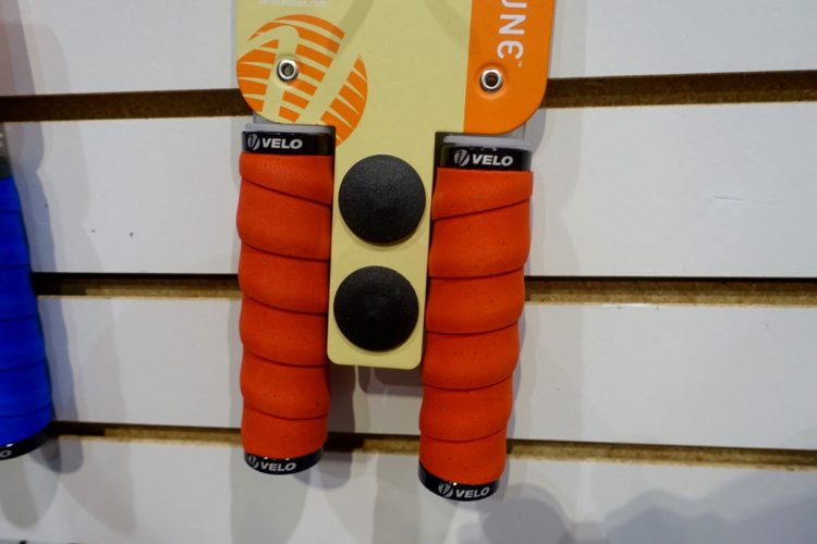 Alt text: Close-up of two orange bicycle handlebar grips with spiral patterns, mounted on a display board. The grips feature the brand name "VELO" at the bottom. Two black rubber end caps are attached between the grips.