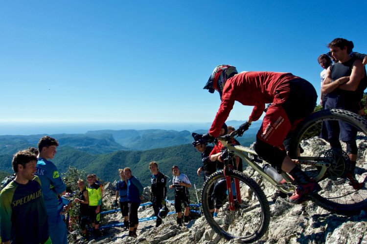 Mountain biker preparing to descend on rocky terrain, surrounded by a group of spectators enjoying the view of a lush valley and hills under a clear blue sky.