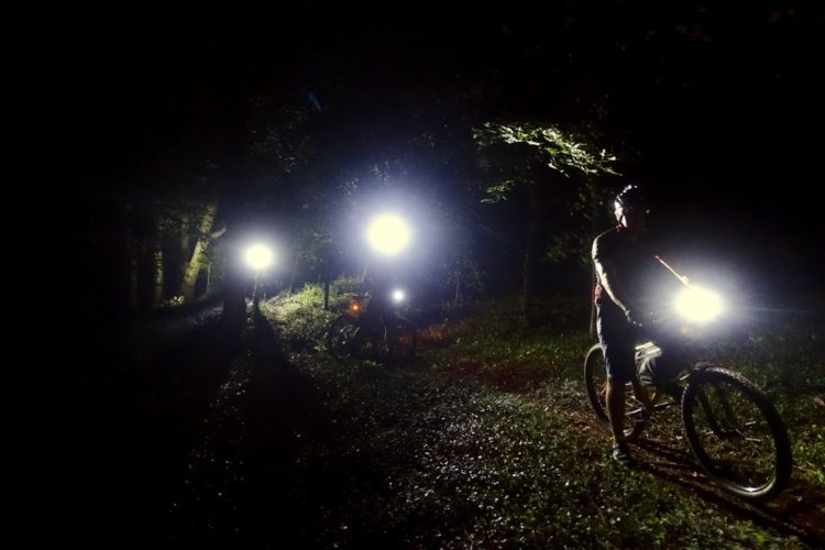 Mountain bikers riding on a dimly lit trail at night, with bright bike lights illuminating the surrounding trees and ground. The scene captures a sense of adventure in a forested environment.