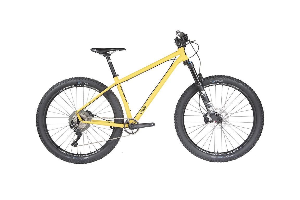 Esker Hayduke: A yellow mountain bike with large black tires and a sleek design, positioned against a white background. The bike features a front suspension fork, modern gearing system, and a comfortable saddle, making it suitable for off-road trails and rugged terrain.
