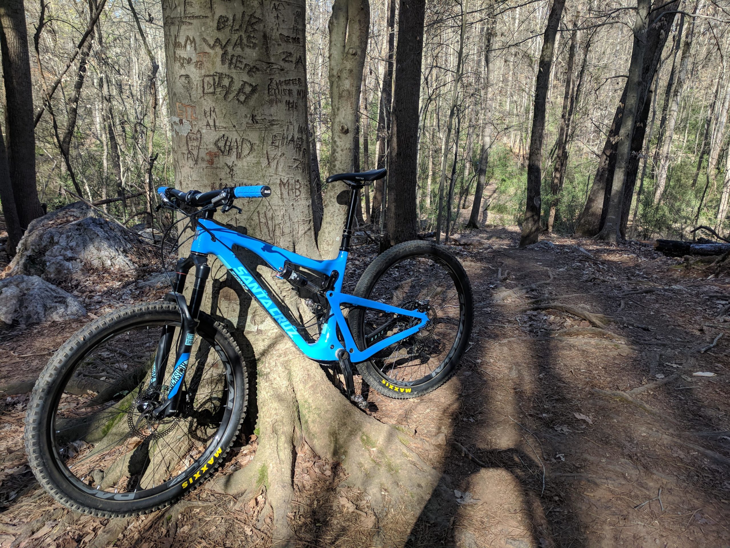 A bright blue mountain bike leaning against a tree in a wooded area, with a dirt trail visible in the background. The tree trunk features carvings and there are rocks and fallen leaves on the ground. USNWC mountain bike trail.