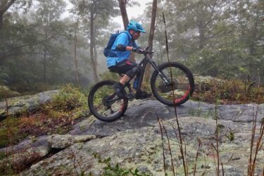 A mountain biker wearing a blue helmet and a blue shirt navigates a rocky trail in a misty forest. The bike's wheels are positioned on a large rock, showcasing the rider's skill in maneuvering through challenging terrain surrounded by greenery and trees. Frederick Watershed mountain bike trail.