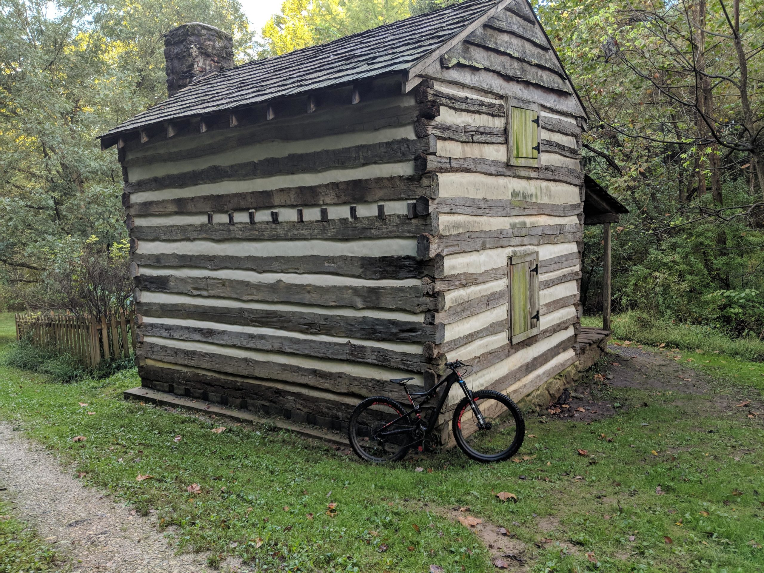 A rustic log cabin with a stone chimney, featuring horizontal wooden beams and bright green window shutters. A mountain bike leans against the side of the cabin, surrounded by lush greenery and a gravel path leading up to it. The scene is set in a wooded area with trees in the background. Hashawha mountain bike trail.