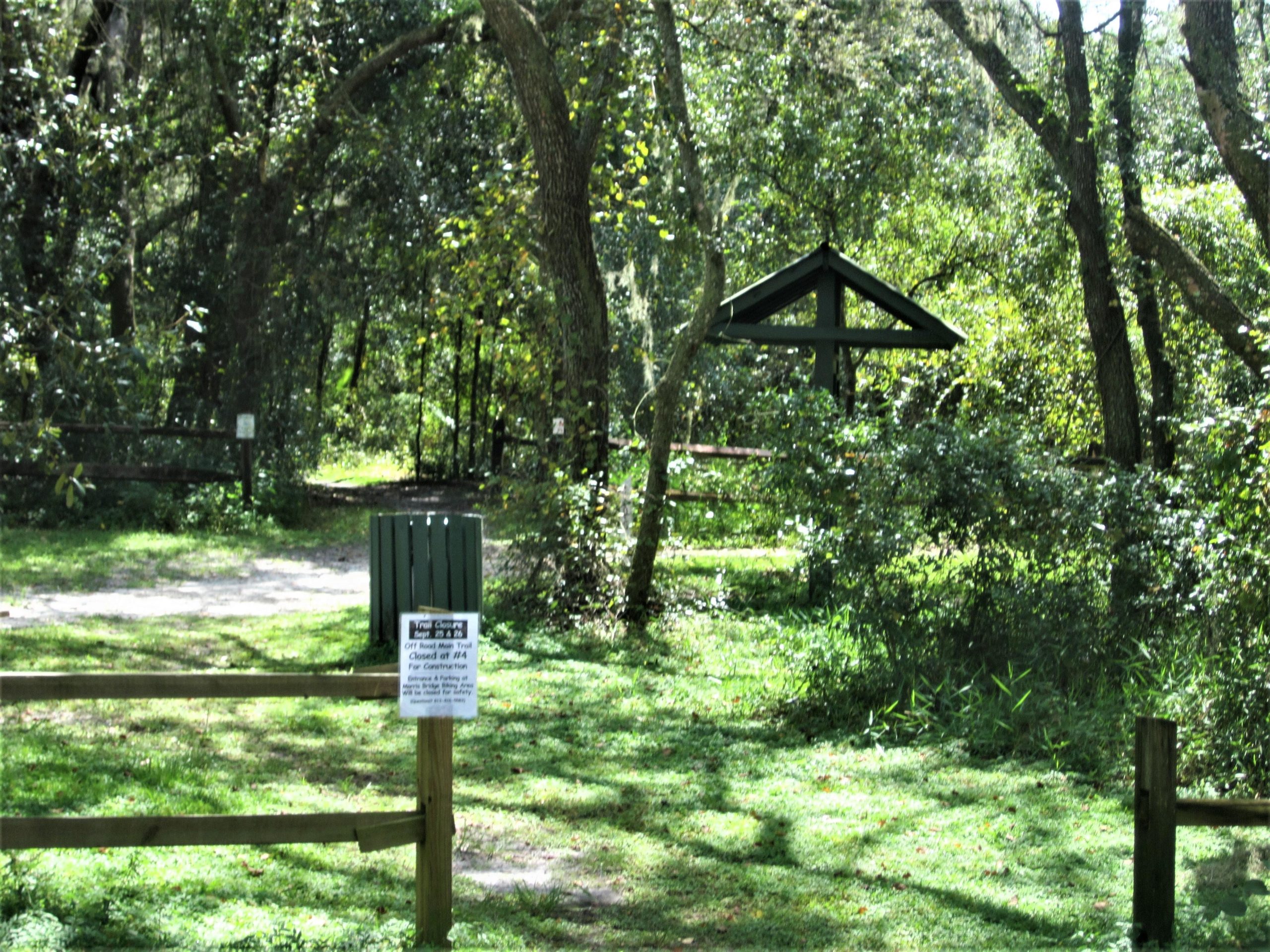A sunlit pathway leads into a lush green forest, bordered by wooden fencing. To the left, there is a wooden post with a sign detailing off-road trail closures. In the background, a shaded shelter with a triangular roof can be seen amidst the trees, surrounded by vibrant foliage. Wilderness Trails Park Loop mountain bike trail.